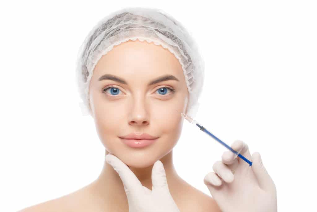 Close-up portrait of young female wearing medical cap, getting ready for filler injection to eliminate aging effect, isolated on white background